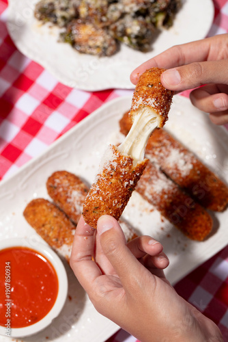 Hands pulling apart a fried cheese stick on a white plate with a red and white checkered tablecloth 