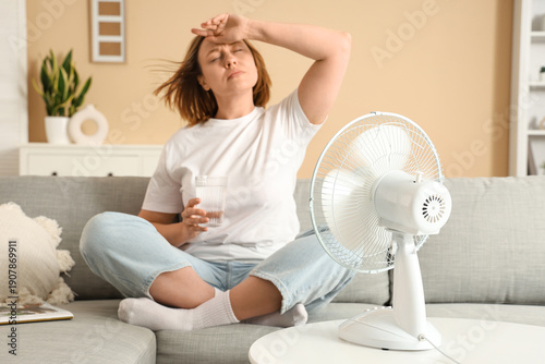 Fotografía Beautiful mature woman with electric fan and glass of water suffering from heat