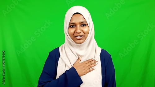 Portrait of a smiling young Muslim woman wearing a hijab with her hand on her heart against a green screen background during ramadan.