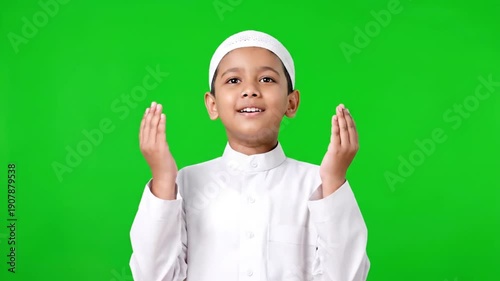 A young Muslim boy wearing a white prayer cap and robe with hands raised in prayer against a green background during ramadan.