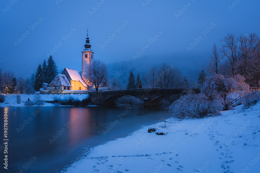 Fototapeta premium Snowy church by Lake Bohinj in Slovenia reflecting calm winter evening atmosphere