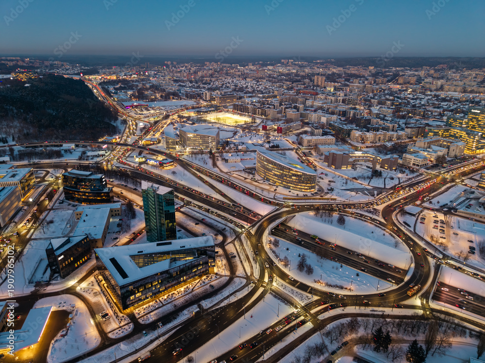 Fototapeta premium Aerial view of a winter sunset over the traffic circle connecting Geležinio Vilko Street and Konstitucijos Avenue in Vilnius, Lithuania. Warm sunset light contrasts with snowy urban infrastructure.