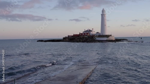 A static medium shot of the historic St Mary’s Lighthouse in Whitley Bay at dawn, featuring the white tower and red-roofed cottages on a tidal island with a rotating offshore wind turbine 