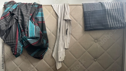 Canvas Print Various clothes, including striped and patterned fabrics, and a plain grey garment, casually draped over a padded, tufted headboard, showing textured textiles