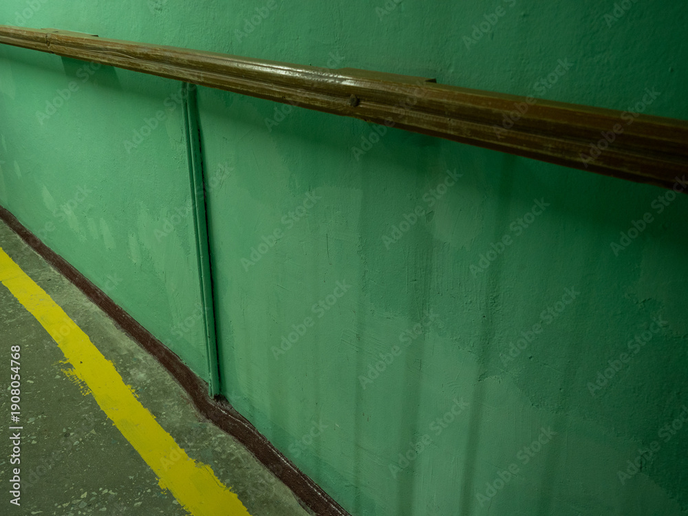 Obraz premium Weathered green textured wall with wooden handrail and yellow safety line painted on concrete floor inside industrial facility or manufacturing plant corridor