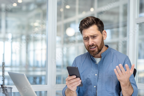 Konstfotografi Young man feeling confused and frustrated, holding a smartphone in an office set