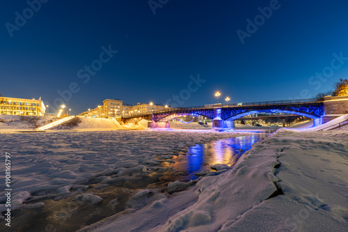Night view of the frozen Neris River in Vilnius with Žvėrynas Bridge illuminated in red and blue lights, the Lithuanian Parliament in the background, and faint stars visible in the winter sky.