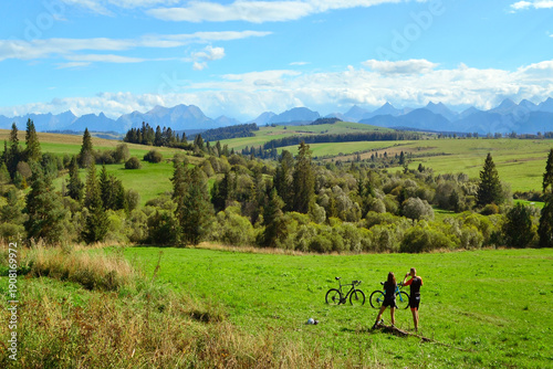 Young couple cyclists, man and woman standing near bicycles on grassy hill  and looking at  mountains. A route for cyclists and hikers around the Tatra Mountains near Trstena village, Slovakia.