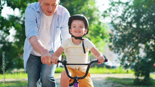 Child wearing helmet father learning to ride bike outdoors in summer. Smiling boy cyclist plays with dad city park. Family weekend. Dad teaches his young son to ride bike in park. Happy family, nature