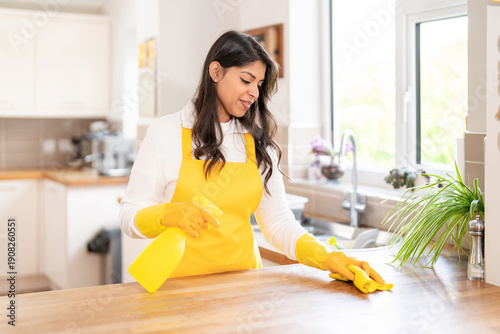 Принти на полотні Woman cleans kitchen counter with spray and cloth while wearing yellow gloves an