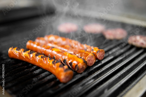 Grilled sausages with spiral cuts and char marks sizzle on a hot metal barbecue grate with rising smoke.