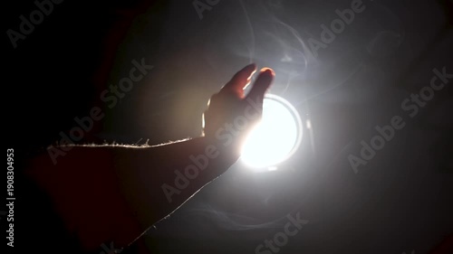 A man's hand stretches out towards a intense beam of light in a hazy atmosphere. Soft-focus close-up symbolizes the search for divine truth and revelation.