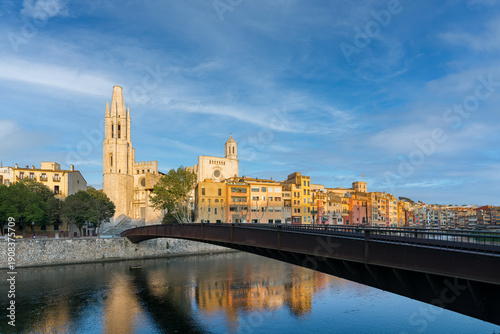 Gironas colorful medieval houses and bridges along the Onyar River, with Sant Feliu church and the cathedral reflected in calm water under a bright blue sky