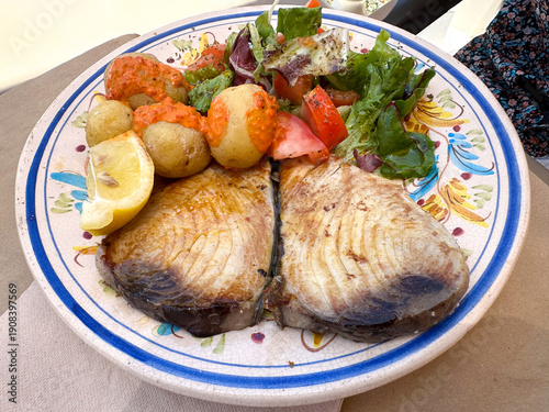 Grilled tuna fish steak served on a decorative patterned plate with fresh vegetables, lemon wedges, and boiled potatoes with salt, showcasing a delicious Canarian traditional culinary presentation