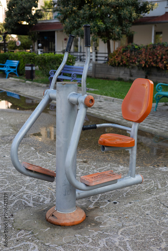 Outdoor fitness machine with a seat designed for strength and cardio exercises, installed in a city park. Metal structure with plastic seating for comfortable outdoor workouts.