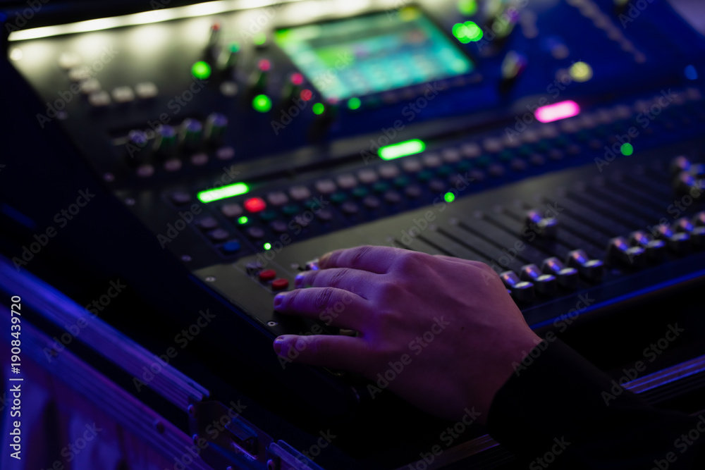 Fototapeta premium Hand on digital audio mixing console in road case, adjusting faders and backlit buttons for live event sound control under blue stage lighting