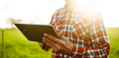 A man stands in a green field at sunset, holding a tablet. The sunlight creates a warm glow. The man is wearing a plaid shirt and appears to be staring intently at the device.