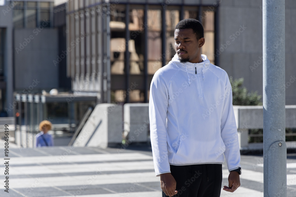 Fototapeta premium African American man standing near metal light pole on tiled plaza wearing white hoodie, copy space