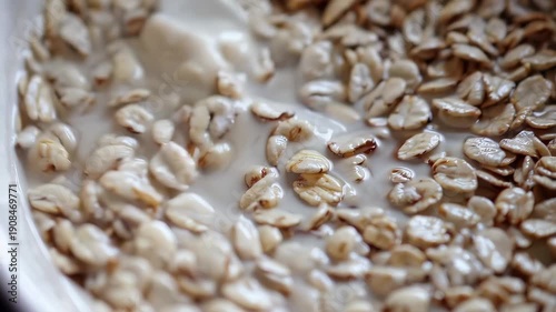 Creamy milk is stirred into a bowl of dry oatmeal flakes with a white spoon. Soft, natural light illuminates the breakfast meal, perfect for a health advertisement.