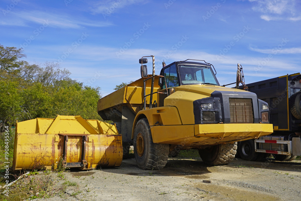 Obraz premium Dump truck with attachment. Yellow articulated dump truck and yellow attachment container parked on gravel near trees and other trucks.