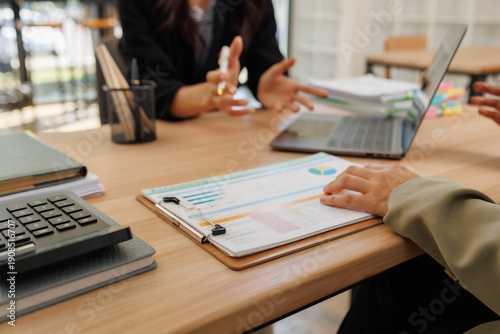 Two people are sitting at a table with papers and a calculator. One of them is holding a piece of paper with a graph on it. The other person is writing in a notebook
