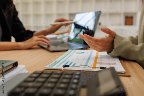 Two people are sitting at a table with papers and a calculator. One of them is holding a piece of paper with a graph on it. The other person is writing in a notebook
