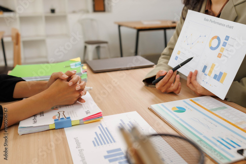 Two people are sitting at a table with papers and a calculator. One of them is holding a piece of paper with a graph on it. The other person is writing in a notebook
