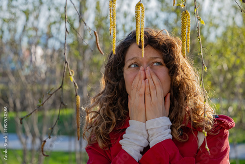 Canvastavla Young woman with curly hair wearing a red coat is covering her nose and mouth wi