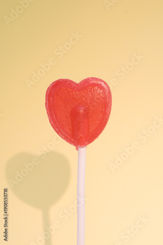 Red heart-shaped lollipop creating a shadow on a soft yellow background