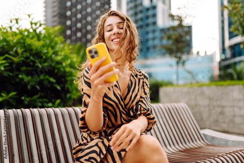 Fotografija A portrait of a happy woman with a phone stands on a city street overlooking a metropolis