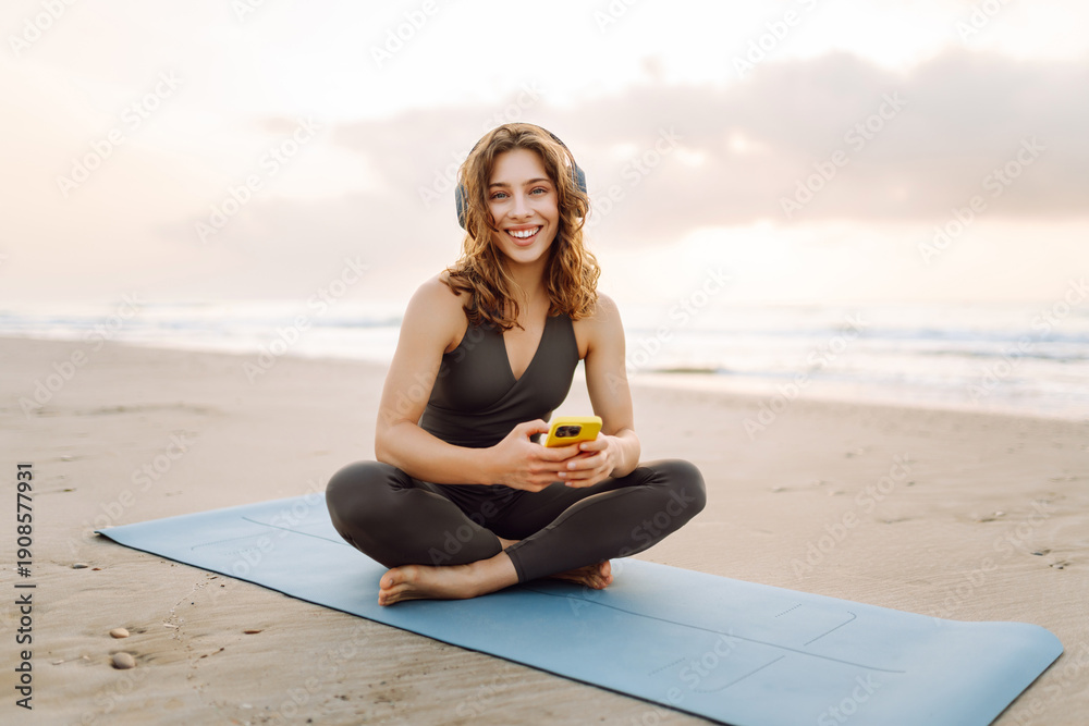 Fototapeta premium Woman on blue mat with phone and wireless headphones against backdrop of sea. Athletic woman listens to music and enjoys sunrise against backdrop of seascape. Concept of meditation and relaxation.
