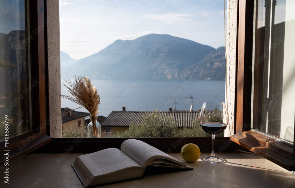 Obraz premium Lake Garda view through window with wine glass and lemons on table. Italian vacation mood, slow living and romantic lifestyle concept.