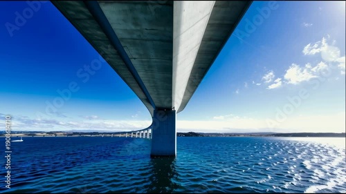 A picturesque view of the long bridge spanning the bay from water level. A cinematic flyover beneath the engineering structure, clear azure water, and an endless horizon.