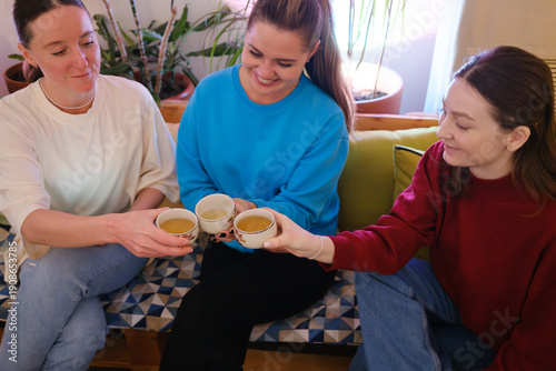 Friends share a cozy tea moment on a sofa, toasting mugs in a warm living room