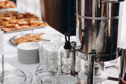Set of coffee break lunch in the hotel restaurant during conference meeting, with tea and coffee catering, decorated banquet table with white cups, tableware and metal tea maker machine boiler