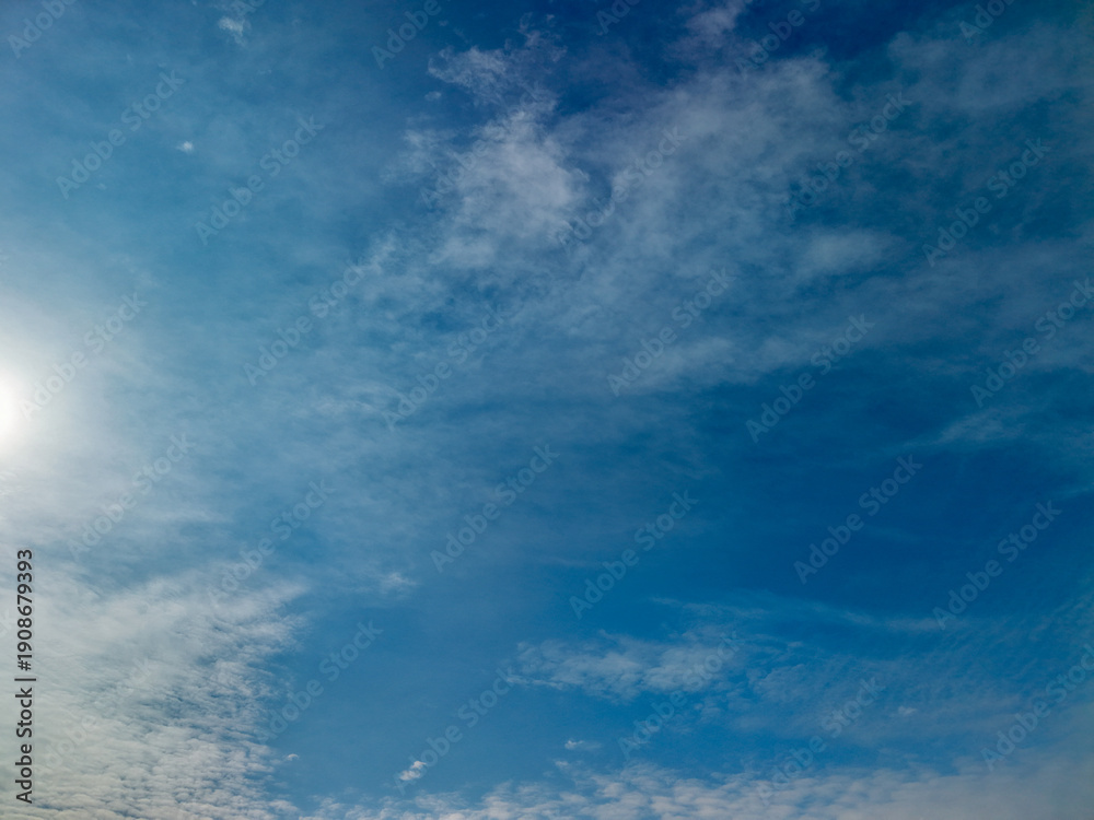 Fototapeta premium Expansive blue sky featuring soft, white wispy clouds, a bright sunburst in the upper left corner, and a clear atmospheric background representing concepts of freedom, nature, and weather