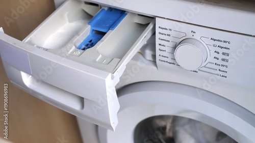 Close-up view of a washing machine detergent drawer with blue compartment and control dial, showcasing settings for various wash cycles and temperature options in a modern laundry room setting