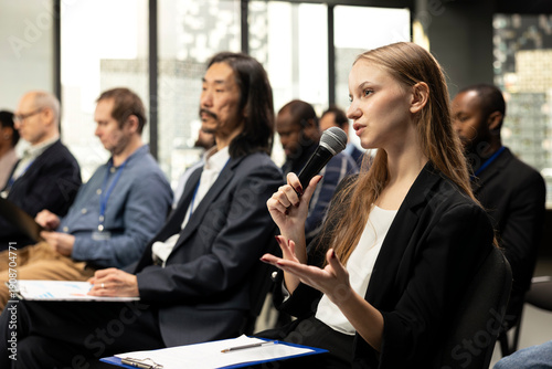Photos Young woman journalist reading questions from clipboard and taking notes, interacting with presenter via microphone in conference room