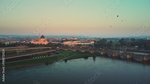 At dawn in Dresden's Old Town, a train crosses the bridge over the Elbe River, with the Yenidze Theater and Heinz-Steyer-Stadion in the background and a bird's-eye view of hot air balloons in the sky
