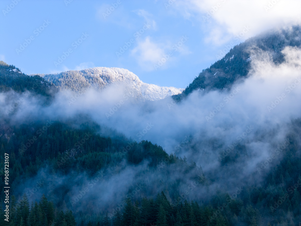 Fototapeta premium Snow-Capped Mountain Ridge Over Misty Forest Valley Under Clear Blue Sky in Howe Sound