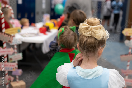 Children in costumes wait in line at a holiday event in a school hallway near festive decorations and tables