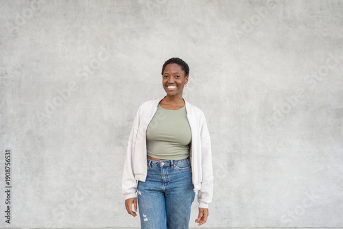Smiling young black woman standing against gray wall wearing trendy casual clothes. Friendly expression with relaxed mood and modern everyday style