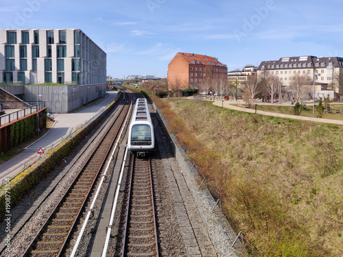 A modern metro train travels along tracks past city buildings and a grassy embankment. Copenhagen.