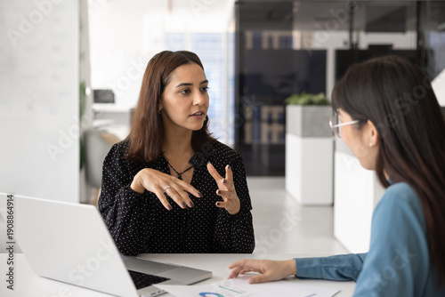 Young Middle Eastern business professional woman talking to consultant, marketing expert, advisor, meeting with colleague at workplace table, discussing analytic report