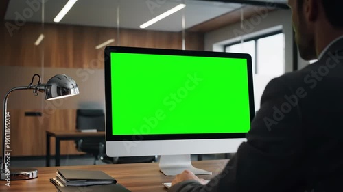 Businessman sitting at wooden desk in modern office with desktop computer showing green screen mockup for screen replacement