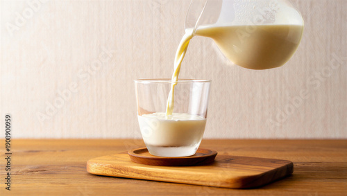 Foto Milk being poured from a glass jug into a clear glass, with a wooden coaster