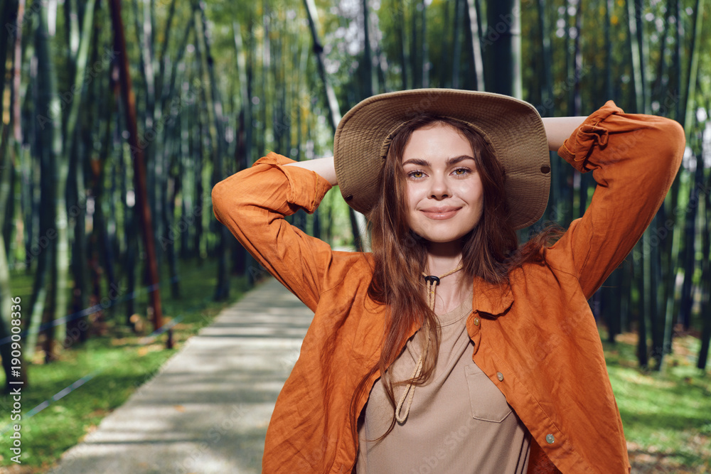 Fototapeta premium Woman smile hat park pathway portrait outdoor fashion lifestyle image of a young woman wearing a wide brim hat and orange jacket standing on a tree lined path, relaxed and confident.