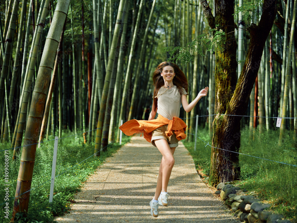 Fototapeta premium Woman in orange skirt jumps with smile on a forest path through tall bamboo, motion and joy captured during outdoor leisure, young model running and twirling on gravel walkway.