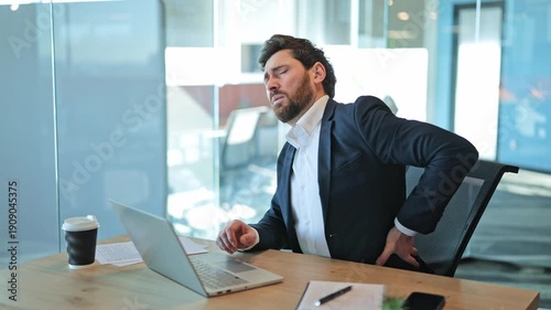 Focused business person feels intense back pain during long hours at office desk. Discomfort strain manifest as chronic posture issues from prolonged laptop use. Suffering and fatigue, seeking relief.