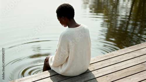 Rear view of a Black woman sitting on a wooden dock by the lake. Person in a white sweater splashing water with feet creating ripples. Solitude and relaxation concept